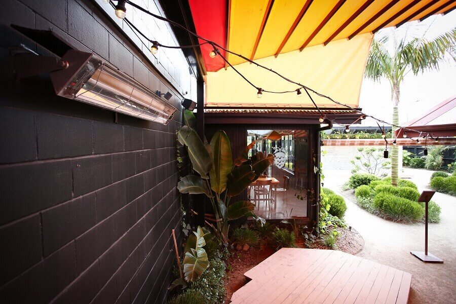 Kelray wall-mounted infrared heater installed in a contemporary café courtyard with black brick walls, festoon lighting, and tropical greenery.