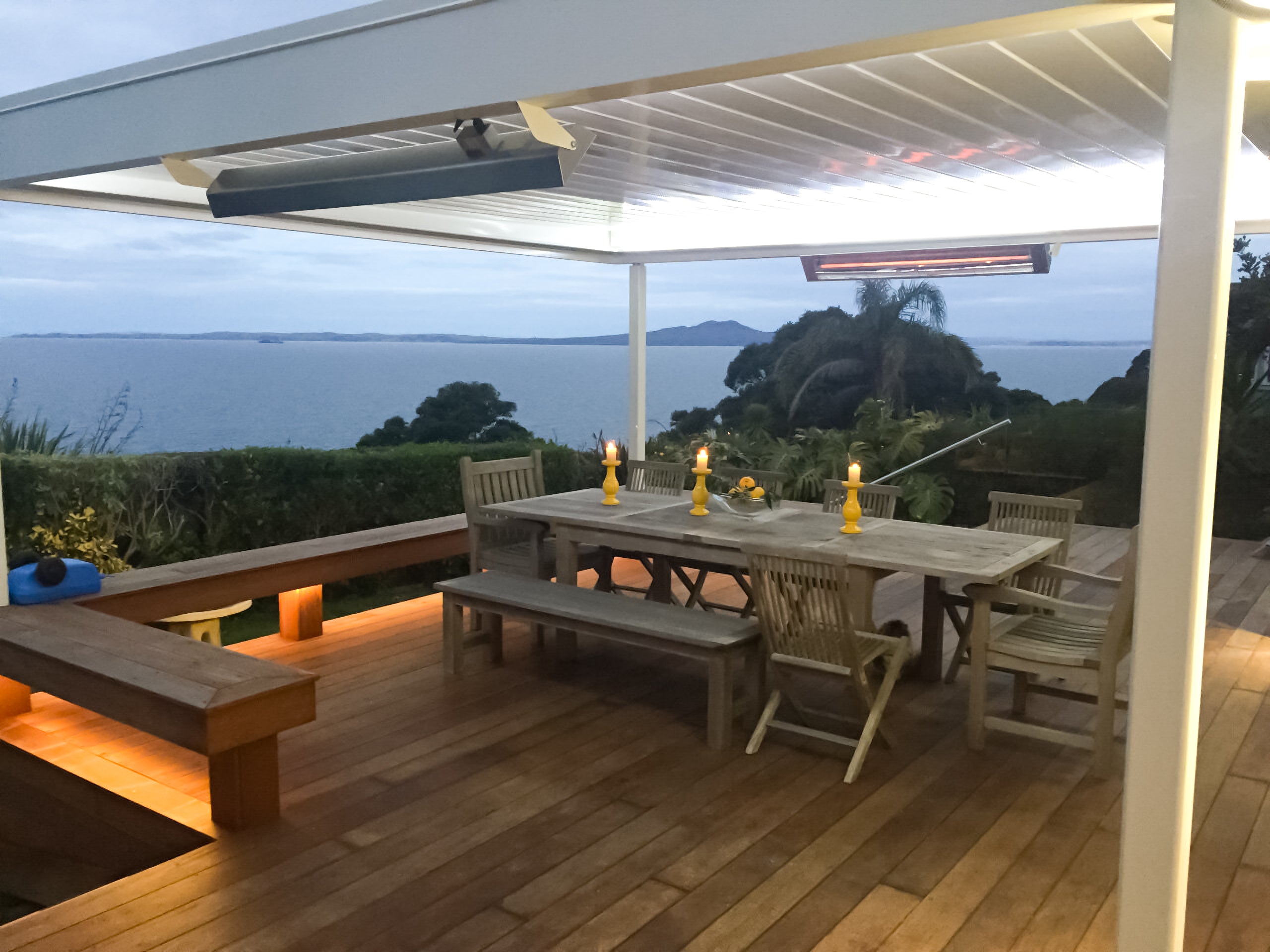 Outdoor dining area with table and chairs on a wooden deck overlooking water and trees with Kelray Infrared Heaters keeping the space warm.