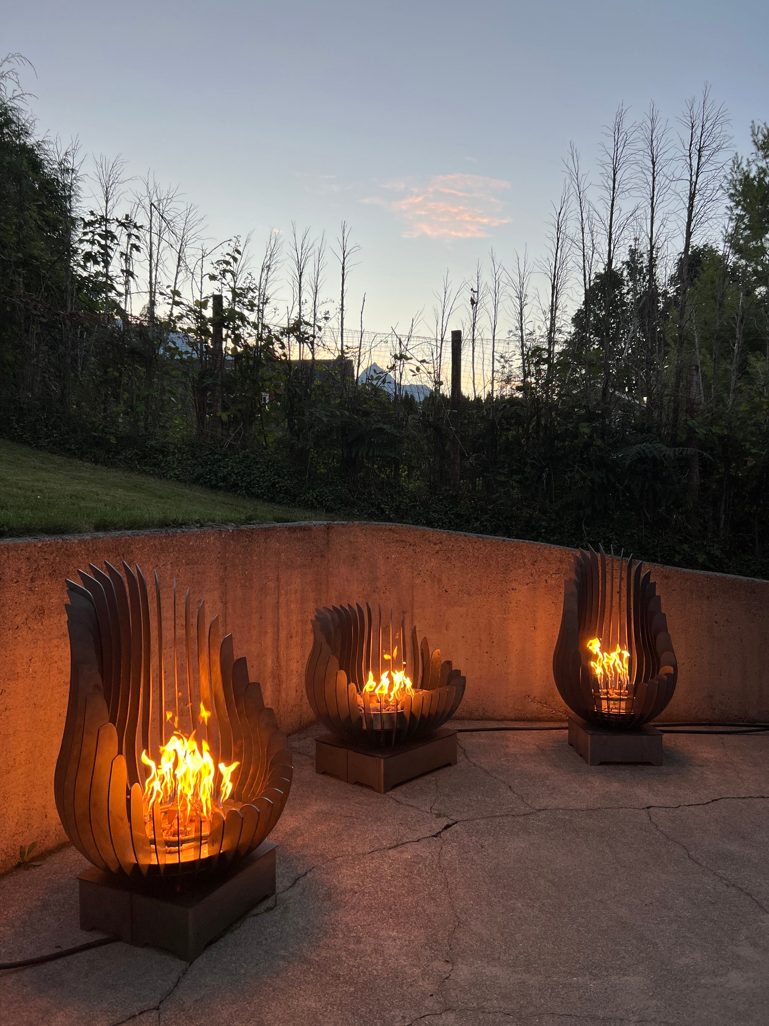 Three Kelray sculptural firepits lit at dusk, arranged in a curved outdoor concrete space with a backdrop of trees and twilight sky.